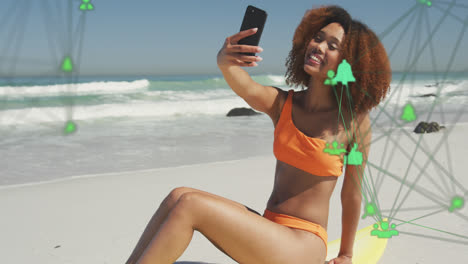 Animation-of-networks-with-digital-icons-over-woman-taking-selfie-with-smartphone-on-beach