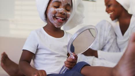 Happy-african-american-mother-and-daughter-sitting-on-bed-daughter-putting-facial-cream