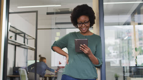 African-american-businesswoman-leaning-in-doorway-using-tablet-smiling-to-camera-in-office