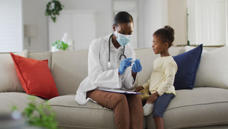African-american-female-doctor-wearing-face-mask-measuring-temperature-of-girl-at-home
