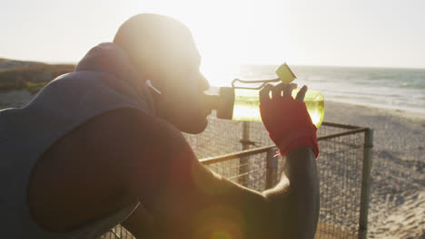 African-american-man-drinking-water,-taking-break-in-exercise-outdoors-by-the-sea