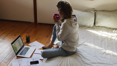 Mixed-race-woman-working-at-home,-sitting-on-bed-using-laptop-and-drinking-tea-in-cottage