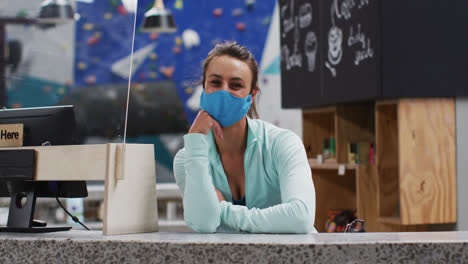 Caucasian-woman-wearing-face-mask-leaning-on-counter-at-reception-of-indoor-climbing-gym