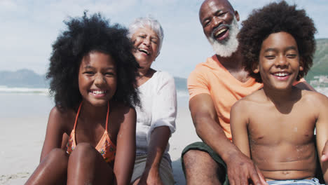 Portrait-of-mixed-race-senior-couple-with-grandchildren-sitting-and-smiling-smiling-at-the-beach