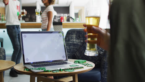 Mixed-race-woman-celebrating-st-patrick's-day-making-video-call-with-laptop-at-a-bar