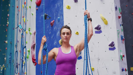Portrait-of-caucasian-woman-wearing-harness-and-holding-ropes-at-indoor-climbing-wall