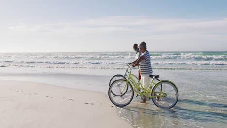 Älteres-Afroamerikanisches-Paar,-Das-Mit-Fahrrädern-Am-Strand-Spazieren-Geht