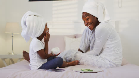 Happy-african-american-mother-and-daughter-wearing-towels-sitting-on-bed-putting-facial-cream