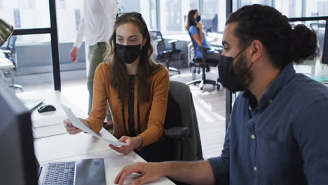 Diverse-male-and-female-colleague-wearing-face-masks-sitting-at-desk-holding-document-in-discussion