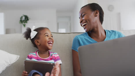 Happy-african-american-mother-and-daughter-sitting-on-sofa-using-digital-tablet,-laptop-and-laughing