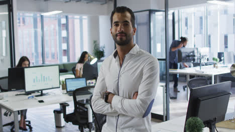 Portrait-of-mixed-race-businessman-standing-in-office-smiling,-with-colleagues-working-in-background