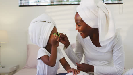 Glückliche-Afroamerikanische-Mutter-Und-Tochter-Tragen-Handtücher-Und-Sitzen-Auf-Dem-Bett,-Legen-Gurken-Auf-Die-Augen