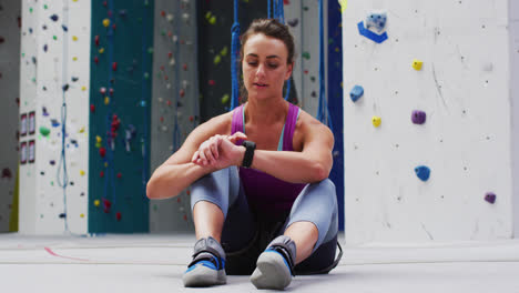 Caucasian-woman-checking-smartwatch-preparing-for-climb-at-indoor-climbing-wall