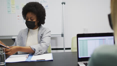 African-american-businesswoman-wearing-face-mask-holding-paperwork-talking-to-colleague-in-office
