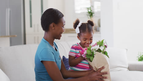 Happy-african-american-mother-and-daughter-sitting-on-sofa-and-smelling-flowers