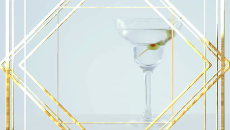 Goldener-Rahmen-Design-Muster-über-Oliven-Im-Cocktailglas-Vor-Grauem-Hintergrund