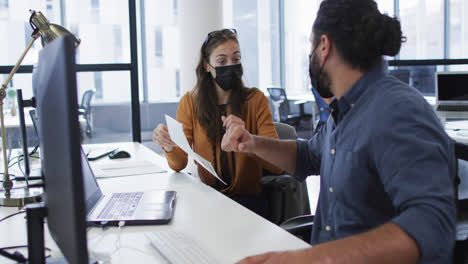 Diverse-male-and-female-colleague-wearing-face-masks-sitting-at-desk-talking-and-elbow-bumping