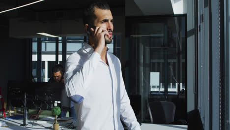 Mixed-race-businessman-standing-in-sunny-office-talking-on-smartphone-and-looking-out-of-window