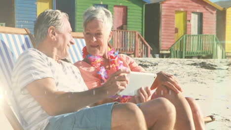 Animation-of-glowing-light-over-portrait-of-happy-senior-couple-using-tablet-in-deckchairs-on-beach