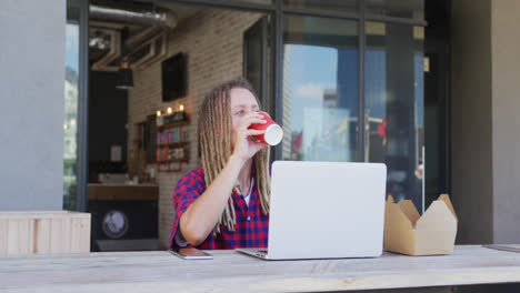 Mixed-race-man-with-dreadlocks-sitting-at-table-outside-cafe-drinking-coffee-and-using-laptop