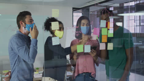 Diverse-colleagues-wearing-face-masks-using-memo-notes-on-glass-wall-having-a-discussion