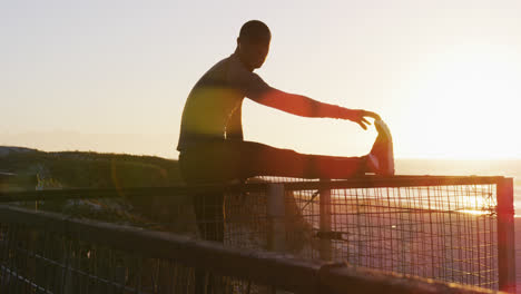Focused-african-american-man-stretching,-exercising-outdoors-by-seaside-at-sunset