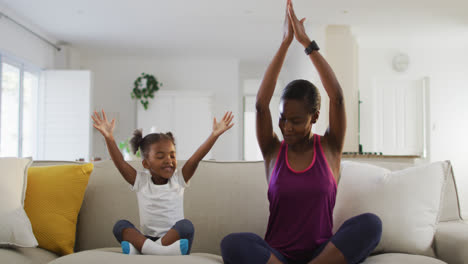Happy-african-american-mother-and-daughter-sitting-on-sofa-doing-yoga-exercise-at-home