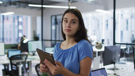 Portrait-of-caucasian-businesswoman-standing-in-office-holding-tablet-and-smiling-to-camera