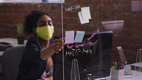 Mixed-race-businesswoman-using-memo-notes-sitting-in-front-of-computer-wearing-face-mask