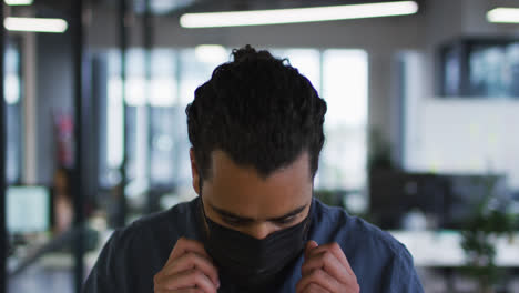 Portrait-of-mixed-race-businessman-adjusting-face-mask-standing-in-office-and-looking-to-camera
