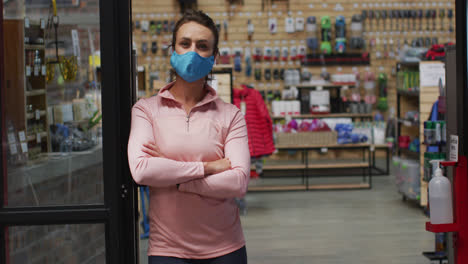 Caucasian-female-shopkeeper-wearing-face-mask-leaning-in-the-doorway-of-sports-shop