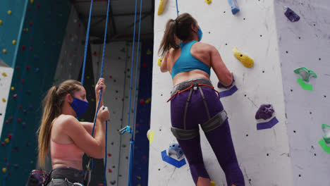 Two-caucasian-women-wearing-face-masks-using-ropes-to-climb-wall-at-indoor-climbing-wall