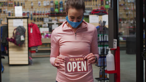 Caucasian-female-shopkeeper-wearing-face-mask-putting-up-open-sign-in-window-of-sports-shop