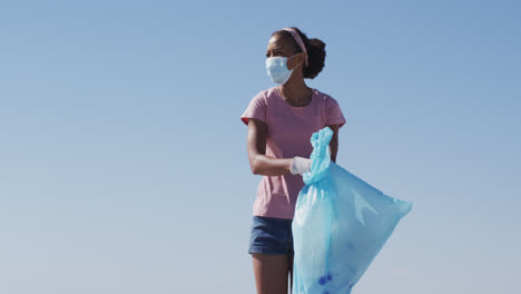 Afroamerikanische-Frau-Mit-Gesichtsmaske,-Die-Einen-Müllsack-Hält-Und-Müll-Vom-Strand-Sammelt