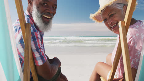 Mixed-race-senior-couple-sitting-on-sunbeds-and-smiling-at-the-beach