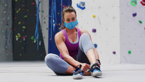 Caucasian-woman-wearing-face-mask-putting-on-climbing-shoes-at-indoor-climbing-wall