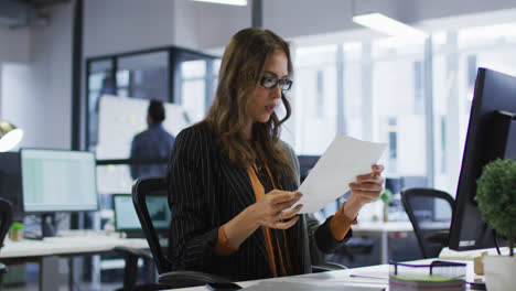 Caucasian-businesswoman-sitting-at-desk-reading-document-and-taking-off-glasses-in-office