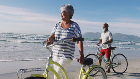 Smiling-senior-african-american-couple-walking-with-bicycles-at-the-beach