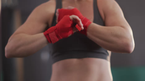 Mid-section-of-caucasian-female-boxer-wrapping-boxing-tape-on-her-hands-at-the-gym