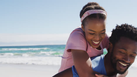African-american-couple-taking-selfie-with-smartphone,-man-carrying-woman-piggyback-on-the-beach
