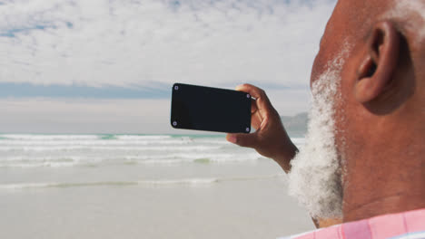 Mixed-race-senior-man-taking-a-selfie-with-a-smartphone-at-the-beach