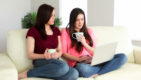 Brunette-using-laptop-and-chatting-to-her-friend-on-the-couch-over-coffee
