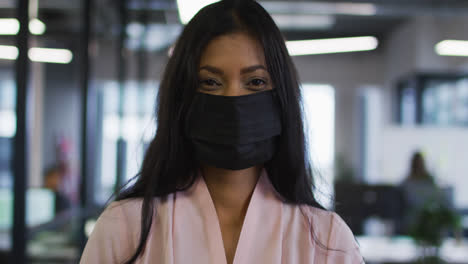 Portrait-of-mixed-race-businesswoman-wearing-face-mask-standing-in-office-looking-to-camera