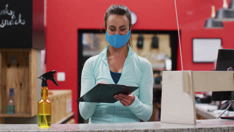 Caucasian-woman-wearing-face-mask-behind-counter-at-reception-of-gym-passing-clipboard