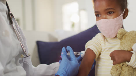 African-american-female-doctor-giving-covid-19-vaccination-to-wearing-face-mask-girl-at-home