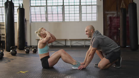 Fit-caucasian-woman-performing-crunches-while-male-trainer-holding-her-legs-at-the-gym