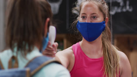 Caucasian-female-gym-receptionist-in-face-mask-taking-temperature-of-female-client