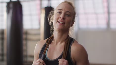 Portrait-of-fit-caucasian-woman-with-jump-ropes-around-her-neck-smiling-at-the-gym