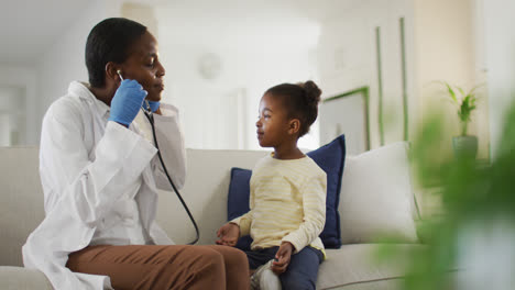 African-american-female-doctor-talking-to-girl-patient-at-home,-using-stethoscope-and-smiling
