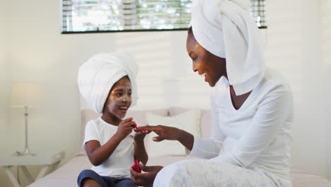 Happy-african-american-mother-and-daughter-sitting-on-bed-and-painting-nails